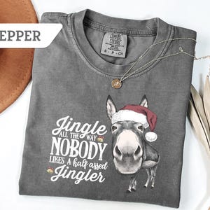 May include: A gray t-shirt with the text "Jingle all the way, nobody likes a half assed Jingler" and a graphic of a donkey wearing a Santa hat. A silver necklace with a pendant is on the shirt.