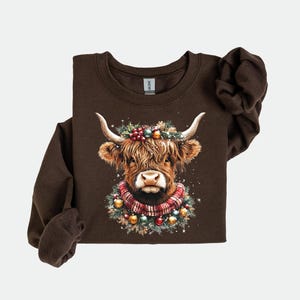 Pu&ograve; includere: Una felpa marrone a maniche lunghe con un'illustrazione ad acquerello di una mucca delle Highlands. La mucca &egrave; adornata con una ghirlanda festiva, bacche, ornamenti e una sciarpa a quadri. Il disegno &egrave; centrato sul petto.