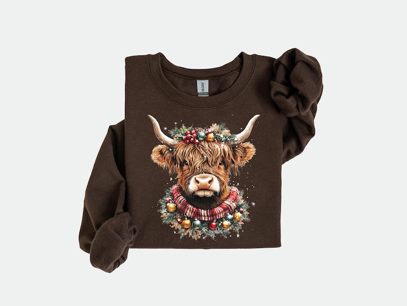 Pu&ograve; includere: Una felpa marrone a maniche lunghe con un'illustrazione ad acquerello di una mucca delle Highlands. La mucca &egrave; adornata con una ghirlanda festiva, bacche, ornamenti e una sciarpa a quadri. Il disegno &egrave; centrato sul petto.