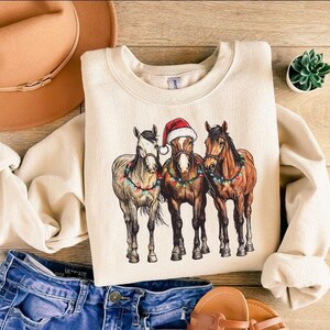 May include: Cream-colored sweatshirt featuring a graphic of three horses adorned with Christmas lights and a Santa hat. The horses are in shades of brown and gray. The sweatshirt is paired with blue jeans, a brown hat, and sandals.