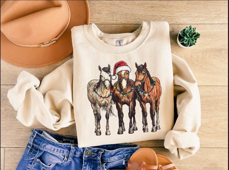 May include: Cream-colored sweatshirt featuring a graphic of three horses adorned with Christmas lights and a Santa hat. The horses are in shades of brown and gray. The sweatshirt is paired with blue jeans, a brown hat, and sandals.