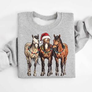 May include: A gray sweatshirt featuring a graphic of three horses. The center horse wears a Santa hat. All three horses are adorned with Christmas lights. The sweatshirt is a crew neck style.