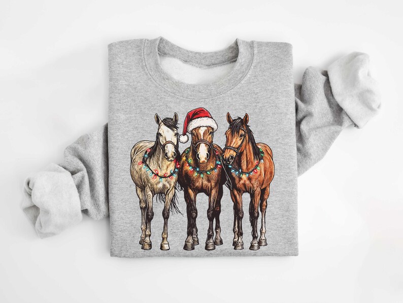 May include: A gray sweatshirt featuring a graphic of three horses. The center horse wears a Santa hat. All three horses are adorned with Christmas lights. The sweatshirt is a crew neck style.