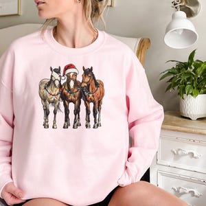 May include: A light pink sweatshirt featuring a graphic of three horses. The center horse wears a Santa hat. The horses are adorned with Christmas lights. The sweatshirt is a crew neck style.
