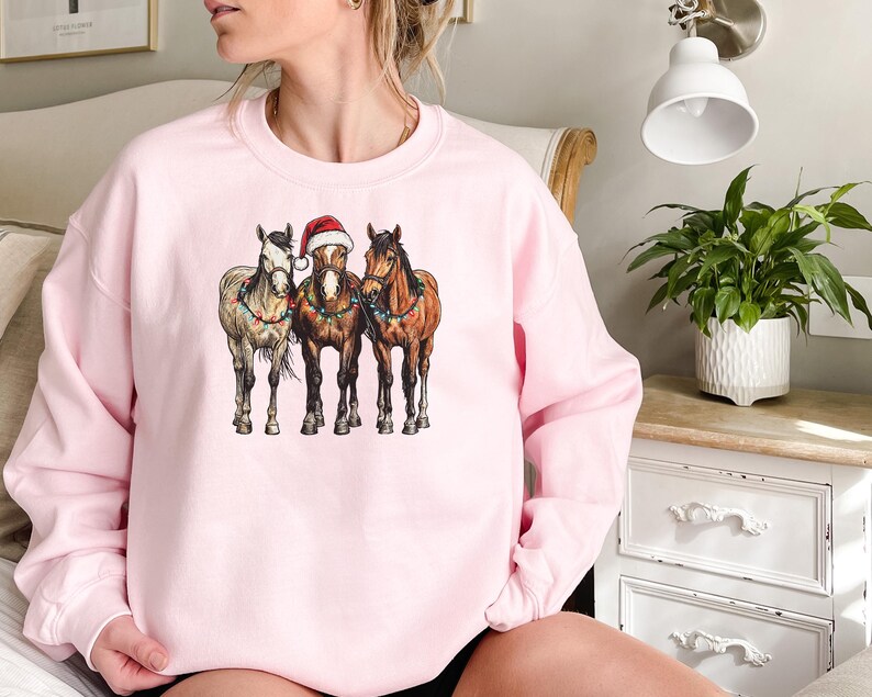 May include: A light pink sweatshirt featuring a graphic of three horses. The center horse wears a Santa hat. The horses are adorned with Christmas lights. The sweatshirt is a crew neck style.