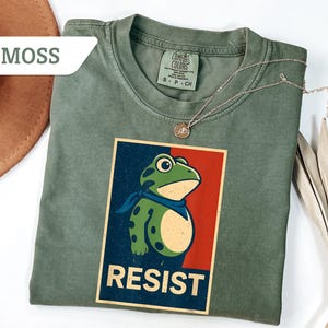 May include: Moss green t-shirt featuring a frog graphic with the word "RESIST" in a vintage poster style. The frog is green with a blue scarf. A gold necklace is draped on the shirt.