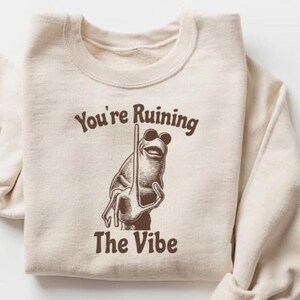 May include: Cream-colored sweatshirt with a brown graphic of a frog wearing sunglasses and holding a pole. The text "You're Ruining The Vibe" is above and below the frog illustration in brown.