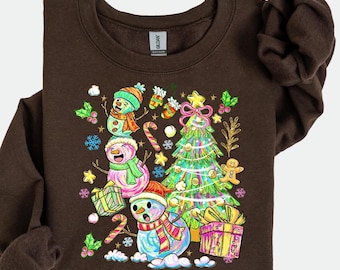 May include: Brown sweatshirt featuring a colorful Christmas design. The graphic includes three snowmen, a decorated Christmas tree, wrapped gifts, candy canes, and snowflakes. The snowmen wear hats and scarves.