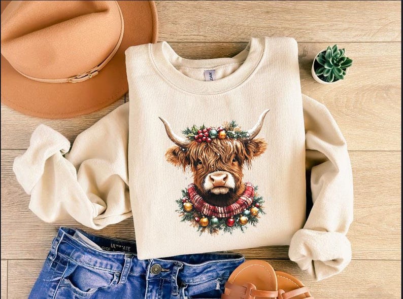 Pu&ograve; includere: Felpa color crema con illustrazione di una mucca delle Highlands con ghirlanda e sciarpa festive. La mucca ha lunghi peli marroni e corna. Il design include ornamenti natalizi. La felpa &egrave; abbinata a jeans blu e un cappello marrone.