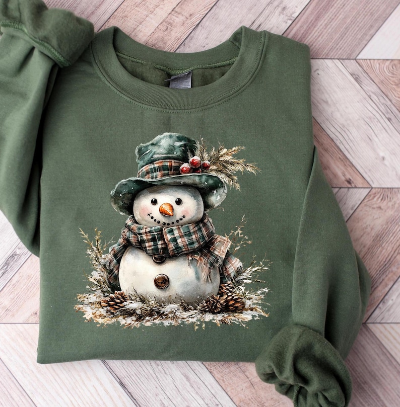 Puede incluir: Sudadera verde con una ilustraci&oacute;n en acuarela de un mu&ntilde;eco de nieve. El mu&ntilde;eco de nieve lleva una bufanda y un sombrero a cuadros, una nariz de zanahoria y ojos de bot&oacute;n. El dise&ntilde;o incluye pi&ntilde;as y ramitas, evocando un tema invernal.