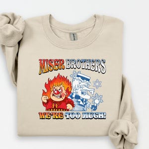 May include: A beige sweatshirt featuring the text "MISER BROTHERS" above a graphic of two cartoon characters, one on fire and the other with snowflakes. The text "WE'RE TOO MUCH!" is below the graphic.