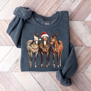 May include: A dark blue sweatshirt featuring a graphic of three horses. The center horse wears a Santa hat. All three horses are adorned with colorful Christmas lights. The sweatshirt is folded on a wooden surface.
