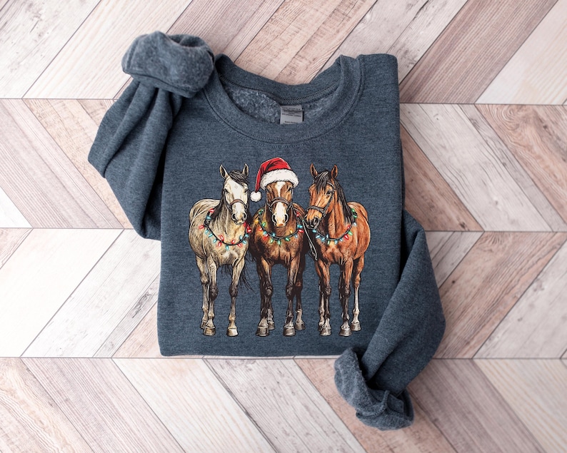 May include: A dark blue sweatshirt featuring a graphic of three horses. The center horse wears a Santa hat. All three horses are adorned with colorful Christmas lights. The sweatshirt is folded on a wooden surface.