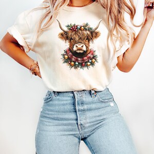 Pu&ograve; includere: T-shirt color crema con grafica di una mucca delle Highlands. La mucca ha lunghi peli marroni, grandi corna e indossa una ghirlanda e una sciarpa natalizia. La maglietta &egrave; abbinata a jeans azzurri.