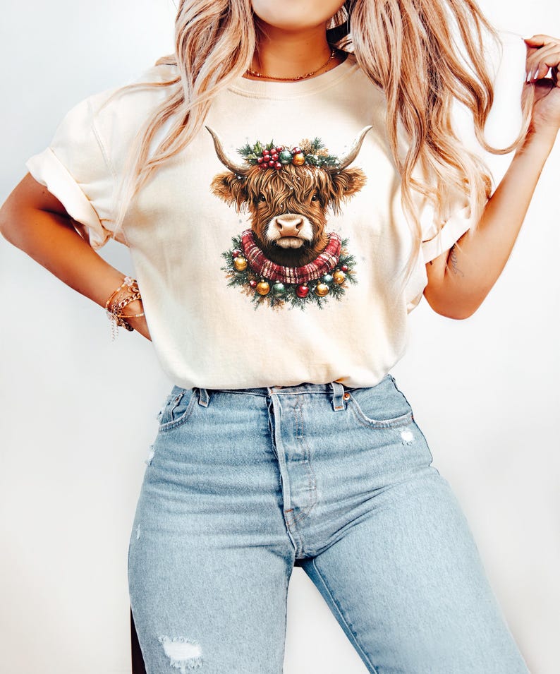 Pu&ograve; includere: T-shirt color crema con grafica di una mucca delle Highlands. La mucca ha lunghi peli marroni, grandi corna e indossa una ghirlanda e una sciarpa natalizia. La maglietta &egrave; abbinata a jeans azzurri.