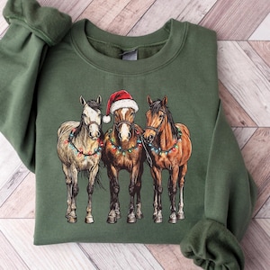 May include: Green sweatshirt featuring a graphic of three horses adorned with Christmas lights. The center horse wears a Santa hat. The horses are brown, tan, and white. The sweatshirt is folded on a wooden surface.