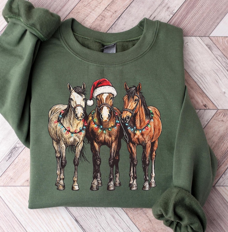 May include: Green sweatshirt featuring a graphic of three horses adorned with Christmas lights. The center horse wears a Santa hat. The horses are brown, tan, and white. The sweatshirt is folded on a wooden surface.
