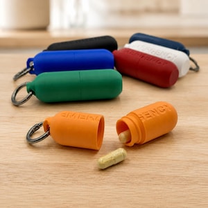 May include: A collection of colorful pill holders with keychains. The holders are in various colors: black, blue, green, red, white, and orange. The orange holder is open, revealing a capsule. The word "EMERGENCY" is printed on the orange holder.