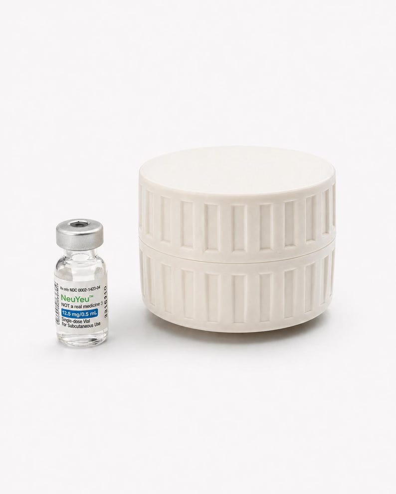 May include: A small glass vial with a silver cap and a white and blue label next to a cream-colored, cylindrical container. The vial contains a clear liquid and the label reads "NeuYeu" and other medical information.