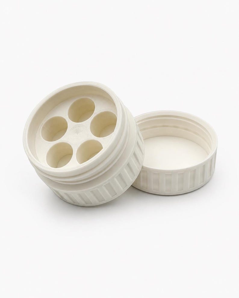 May include: Off-white plastic container with a lid. The container has a circular design with six evenly spaced holes. The lid is separate and has a ribbed texture. The container is designed to hold small items.