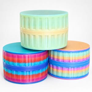May include: Three colorful, cylindrical containers stacked on top of each other. The containers have a ribbed design and are in shades of blue, pink, orange, yellow, and green. The lids are solid colors, with a blue, green, and orange lid.