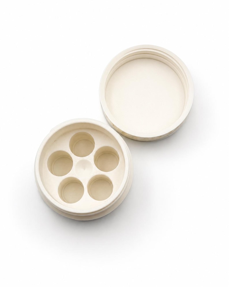 May include: Off-white, circular container with a lid, revealing five small, evenly spaced compartments. The lid is separate, showing the interior. The container is made of a smooth, matte material, likely ceramic or a similar substance.