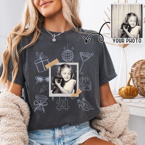 May include: Dark grey t-shirt featuring a framed black and white photo of a girl with a puppy, with light blue illustrations of party items. The number "21" is printed below the photo. The words "YOUR PHOTO" are printed in the upper right corner.