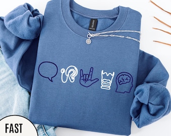 May include: A blue crewneck sweatshirt with white and navy blue graphic icons representing communication, hearing, sign language, and the brain. A silver necklace with a pendant is on the sweatshirt. The sweatshirt is displayed with a pair of blue jeans.