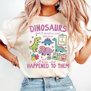 May include: A cream-colored t-shirt featuring a graphic with three colorful dinosaurs, puzzle pieces, and educational charts. The text reads: DINOSAURS didn't reinforce good behavior and we all know what HAPPENED TO THEM, with icons for FIRST THEN and WORK BREAK PLAY.