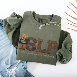 May include: An olive green sweatshirt with the embroidered letters "SLP" in brown, and a teddy bear wearing a "Speech Therapy" sweater. The words "SPEECH LANGUAGE PATHOLOGIST" are embroidered below. A silver necklace is draped across the sweatshirt.