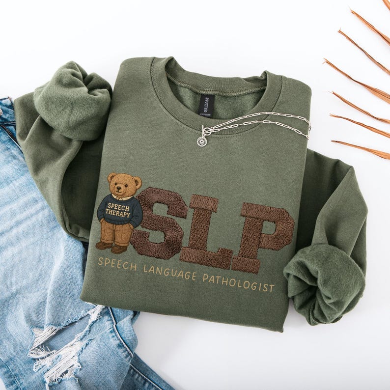 May include: An olive green sweatshirt with the embroidered letters "SLP" in brown, and a teddy bear wearing a "Speech Therapy" sweater. The words "SPEECH LANGUAGE PATHOLOGIST" are embroidered below. A silver necklace is draped across the sweatshirt.