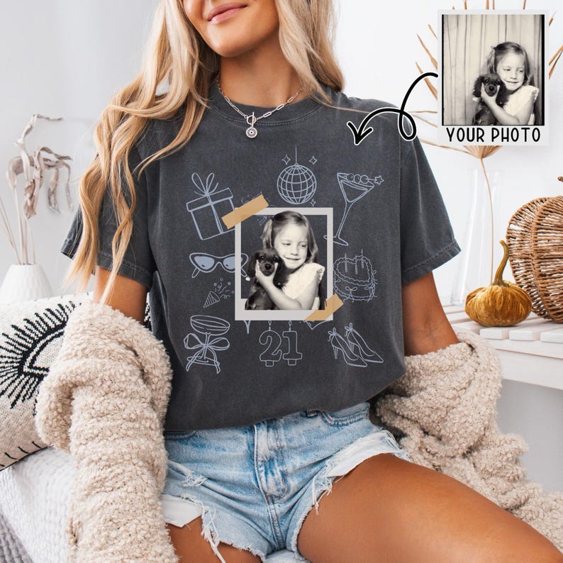 May include: Dark gray t-shirt with a photo of a young girl holding a puppy, surrounded by light blue illustrations of party-themed objects. The number "21" is printed below the photo. The words "YOUR PHOTO" are printed in the upper right corner.
