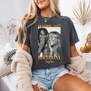 May include: Dark gray t-shirt with a black and white photo of two smiling women. The shirt has gold text that reads "HAPPY 21ST BIRTHDAY Taylor."