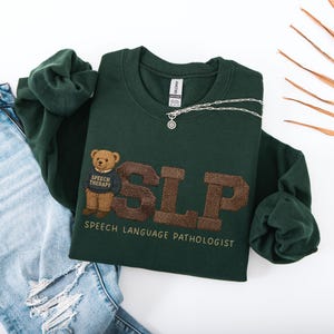 May include: A dark green sweatshirt with the letters "SLP" in brown, embroidered with a teddy bear wearing a "Speech Therapy" shirt. The words "Speech Language Pathologist" are below the letters. A silver necklace is on top.
