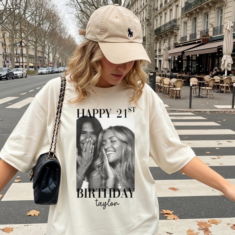 May include: A cream-colored t-shirt with the text "HAPPY 21ST BIRTHDAY taylor" and a black and white photo of two smiling people. The person is wearing a tan baseball cap and carrying a black purse with a chain strap.