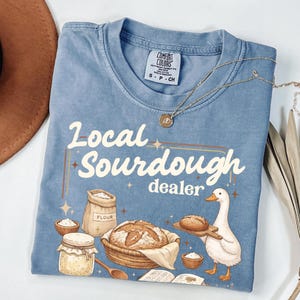Peut inclure: Un t-shirt bleu clair avec le texte "Local Sourdough dealer" et des illustrations de pain, de farine et d'un canard. Un collier doré et un chapeau marron sont également visibles.