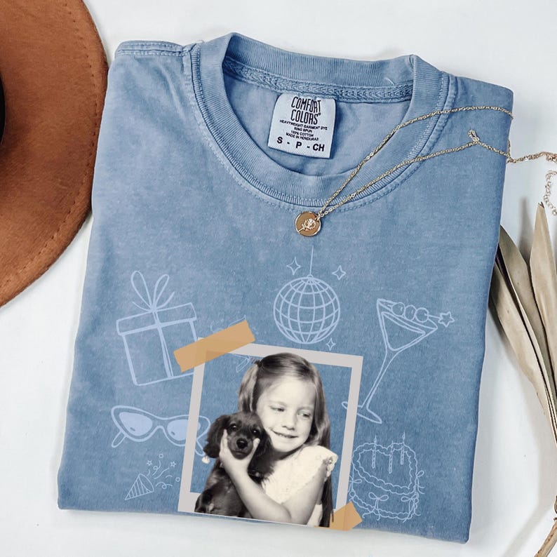 May include: A light blue Comfort Colors t-shirt with a photo of a girl holding a puppy, and party-themed graphics. A gold necklace and a brown hat are also visible. The shirt has the size S-P-CH on the tag.