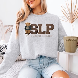 May include: A light gray sweatshirt with the letters "SLP" in brown, with the words "SPEECH LANGUAGE PATHOLOGIST" below. A teddy bear wearing a "SPEECH THERAPY" shirt is on the left. The person is holding a coffee mug.