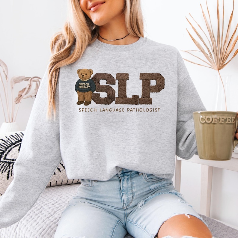 May include: A light gray sweatshirt with the letters "SLP" in brown, with the words "SPEECH LANGUAGE PATHOLOGIST" below. A teddy bear wearing a "SPEECH THERAPY" shirt is on the left. The person is holding a coffee mug.