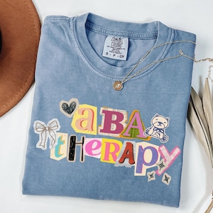 May include: A blue Comfort Colors t-shirt with the words "a BA therapy" in a colorful, collage-style design. The shirt features a small teddy bear graphic and a gold necklace. A brown hat is partially visible in the upper left corner.