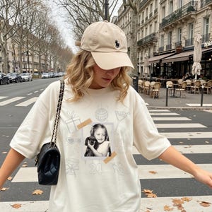 May include: A beige baseball cap with a black logo and a cream-colored t-shirt. The t-shirt features a black and white photo of a girl holding a dog, surrounded by sketched party-themed graphics and the number "21". A black quilted shoulder bag is also visible.
