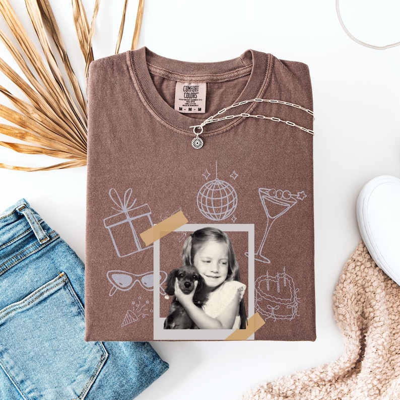 May include: A brown Comfort Colors t-shirt with a graphic of a girl holding a dog, a disco ball, and other party-themed illustrations. A silver necklace with a sun pendant is on the shirt. The shirt is folded on a white surface with a pair of jeans and a shoe.
