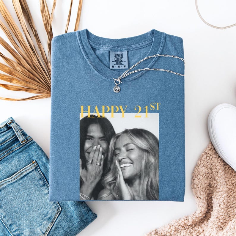 May include: A blue Comfort Colors t-shirt with the text "HAPPY 21ST" and a black and white photo of two women laughing. A silver necklace and a pair of blue jeans are also visible.