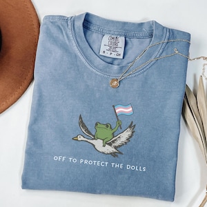 May include: A blue Comfort Colors t-shirt with a graphic of a green frog riding a white goose and holding a transgender pride flag. The text "OFF TO PROTECT THE DOLLS" is printed below the graphic. A gold necklace is draped across the shirt.