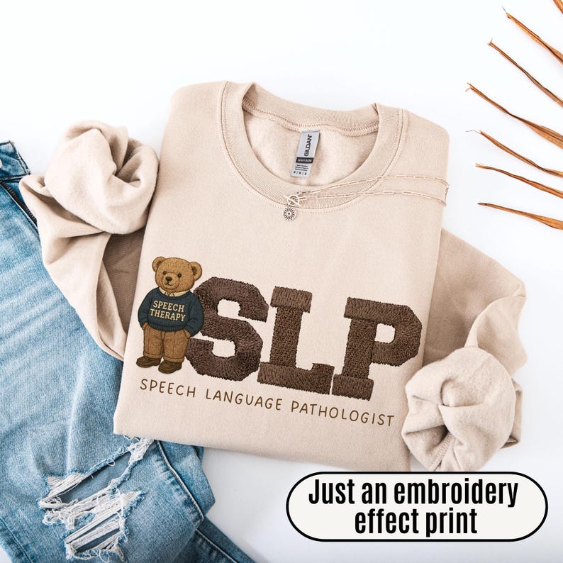 May include: Beige sweatshirt with the letters "SLP" embroidered in brown, along with a teddy bear wearing a "Speech Therapy" sweater. The words "SPEECH LANGUAGE PATHOLOGIST" are below the letters. The text "Just an embroidery effect print" is in a white oval.