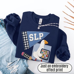 May include: A navy blue sweatshirt featuring a light blue pennant with the letters SLP, a black and white checkered pencil, and a white goose holding a colorful grid of icons. The design includes the text Just an embroidery effect print.