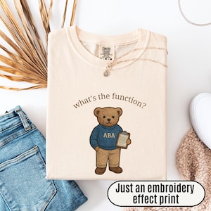 Peut inclure: T-shirt crème avec un dessin d'ours en peluche portant un pull bleu avec "ABA" dessus, tenant un presse-papiers. Le texte "what's the function?" est au-dessus de l'ours. La phrase "Just an embroidery effect print" est en dessous.
