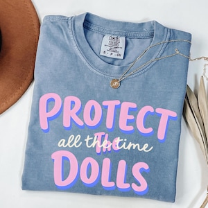May include: A blue Comfort Colors t-shirt with the words "PROTECT all the time DOLLS" in pink and blue lettering. A gold necklace with a pendant is draped across the shirt. A brown hat is visible in the background.