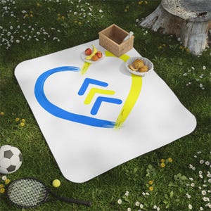 May include: A white picnic blanket with a blue and yellow heart design. The blanket is on a grassy surface with a soccer ball, tennis racket, and a basket of fruit.