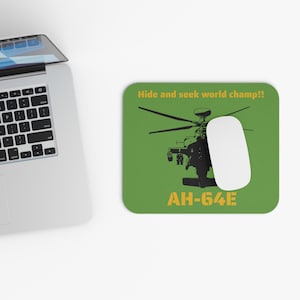 May include: A green rectangular mousepad with a black helicopter graphic and the text "Hide and seek world champ!!" and "AH-64E" in yellow. A white mouse is on the pad, and a laptop is in the background.